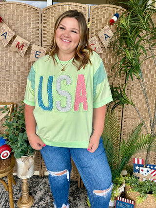 Lime green oversized tee with colorful USA graphic and striped sleeves, paired with distressed denim jeans.
