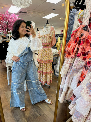 Blue denim overalls with ivory eyelet long-sleeve top worn by woman in store mirror.
