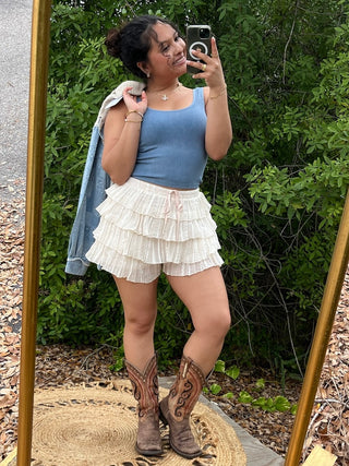 Ivory pleated tiered ruffle shorts with drawstring waist displayed on model with blue ribbed tank top.
