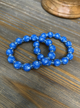 Pair of blue stretch bracelets with white floral pattern beads on wooden surface.
