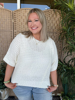 Cream knit sweater with textured weave pattern and short sleeves, loose asymmetrical fit.
