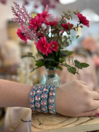 Blue stretch bracelet with pink rhinestones worn on wrist next to magenta flowers.
