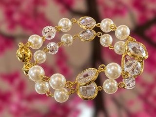 Gold-tone bracelet with cream pearls and faceted crystal links against blurred pink floral background.
