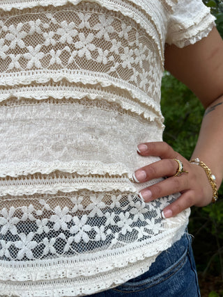 Cream lace crop top with floral and geometric lace panels and ruffled trim details.
