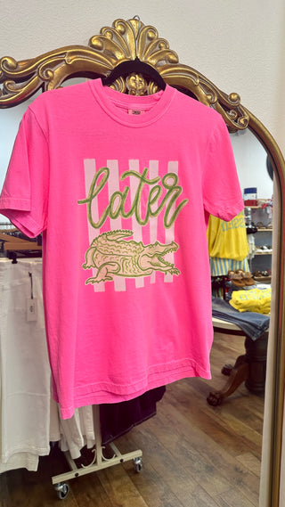 Neon pink crewneck tee with gold "later gator" text and crocodile graphic on checkered background.
