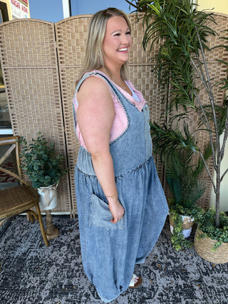 Blue denim sleeveless jumpsuit with pink shirt underneath, oversized fit with front pockets.
