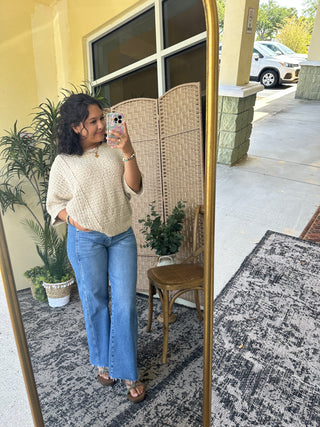 Cream cable-knit asymmetrical sweater top with textured weave, worn with blue jeans.
