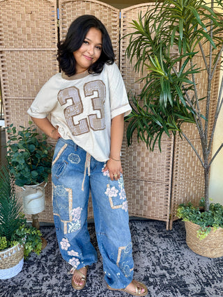 Cream jersey tee with mocha "23" graphic, paired with distressed blue jeans.
