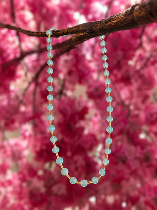 Turquoise glass bead necklace with gold spacers hanging from brown branch against pink floral background.
