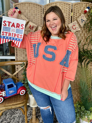 Coral oversized pullover with USA graphic and striped sleeves, paired with distressed blue jeans.
