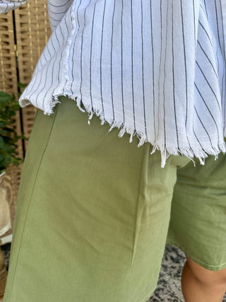 White pinstriped boho shirt with distressed frayed hem worn over sage green pants.
