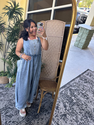 Light blue denim sleeveless jumpsuit with button front and oversized fit, worn with white slide sandals.
