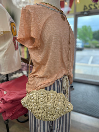 Beige crocheted shell-shaped crossbody bag with woven strap displayed on mannequin wearing peach linen top.
