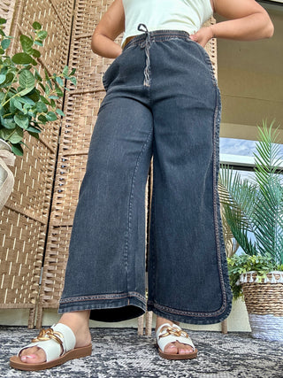 Dark wash wide-leg denim pants with decorative stitching detail, worn with white tank top and white slide sandals.
