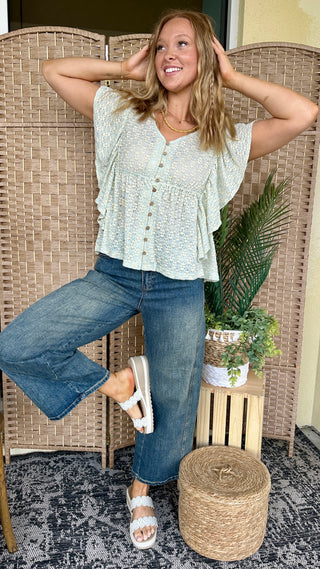 Sage green textured short-sleeve top with button front and tie back detail paired with blue jeans.
