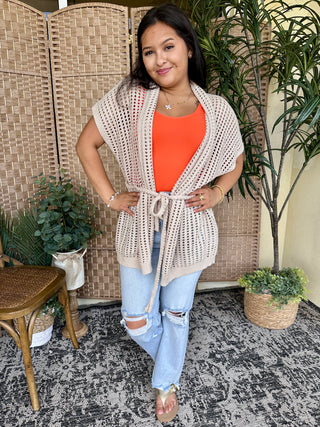 Cream open weave cardigan with tie waist worn over orange tee and distressed light wash jeans.

