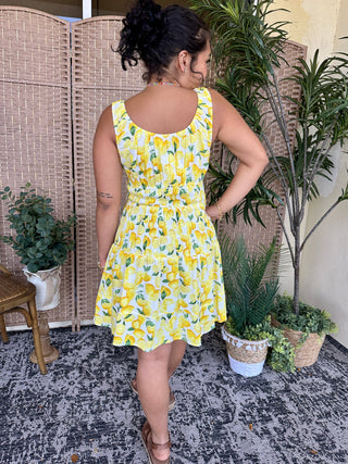 Yellow lemon print mini dress with gathered waist and ruffle neckline on cream background.
