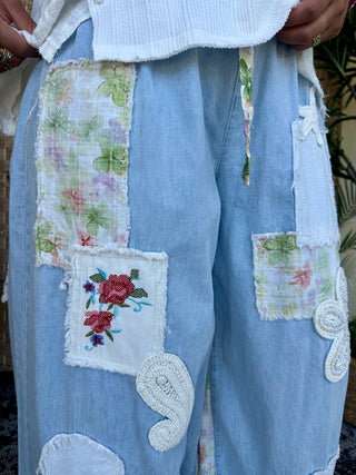 Light blue denim joggers with embroidered floral patches, white crochet details, and distressed edges.
