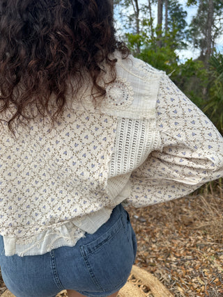 Cream floral jacket with blue ditsy print, lace details, and embellished button on shoulder.
