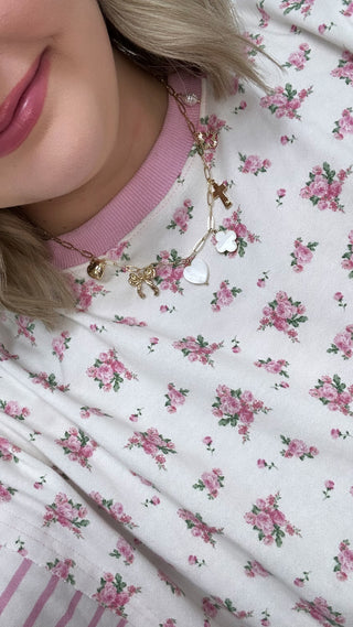 Gold charm necklace with heart, cross, bow and clover pendants on pink ribbed sweater over pink floral print fabric.
