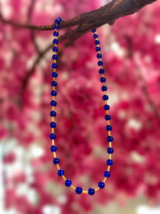 Cobalt blue glass bead necklace with gold spacers hanging from branch with pink floral background.
