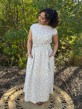 Ivory floral sleeveless midi dress with blue pattern and pleated waist, featuring boat neckline and pockets.
