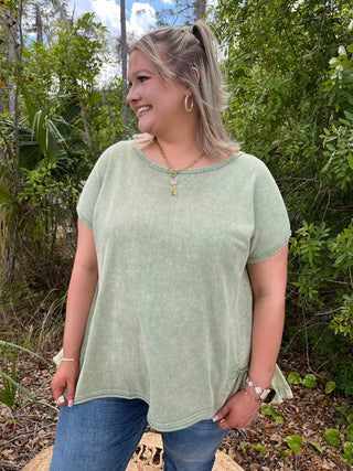 Sage green short-sleeve top with ruffle trim neckline and hem, worn with blue jeans.
