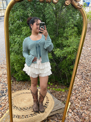 Sage green open-front cardigan with tie closure and openwork knit texture, styled with cream tiered pleated skirt.

