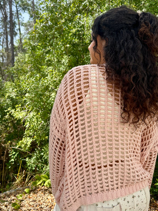 Blush pink open-weave crochet cardigan with geometric cutout pattern worn over white top.
