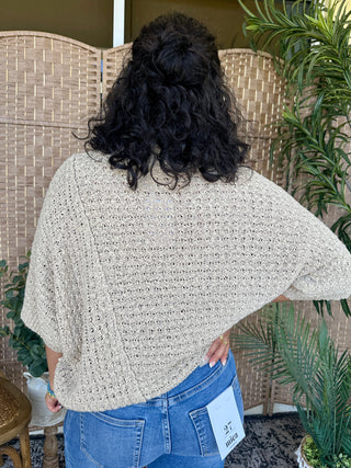 Taupe open knit asymmetrical sweater top worn over blue jeans, showing draped oversized fit.
