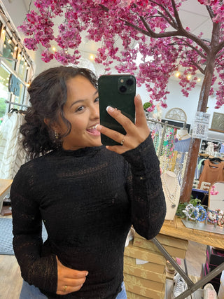 Black textured lace long-sleeve top with sheer sleeves on model in boutique setting.
