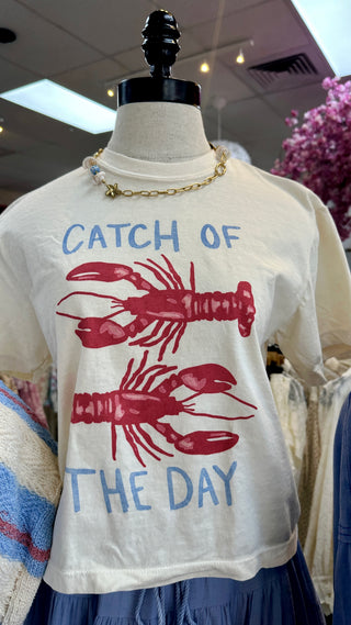 Cream crop tee with blue "Catch of the Day" text and red lobster graphics.
