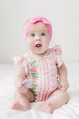 Pink bodysuit with floral rose print and bow pattern, ruffled sleeves, on baby girl with pink headwrap.
