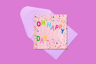 Colorful "Oh Happy Day" greeting card with confetti pattern on pink envelope against purple background.
