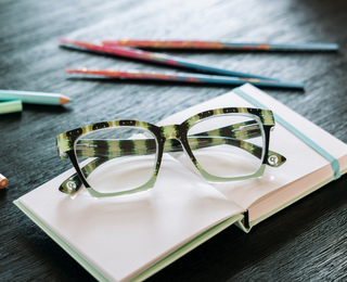 Tortoiseshell and green square-frame reading glasses on open notebook with colored pencils.

