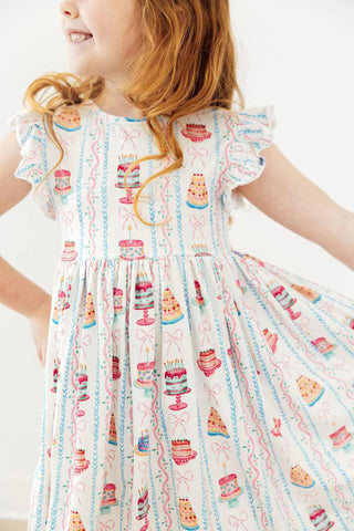 Girls flutter sleeve dress with cake and bow print on cream background.
